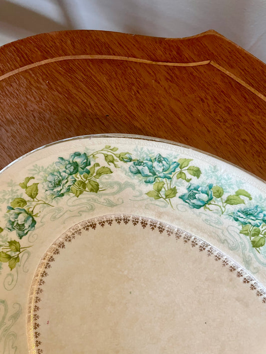 A close-up of an off-white vintage porcelain plate with a floral border design in teal and green, resting on a wooden surface.
