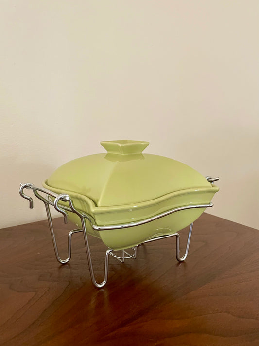 An avocado green ceramic chafing dish with a matching lid, resting on a silver metal wire stand on a wooden table against a plain beige wall.