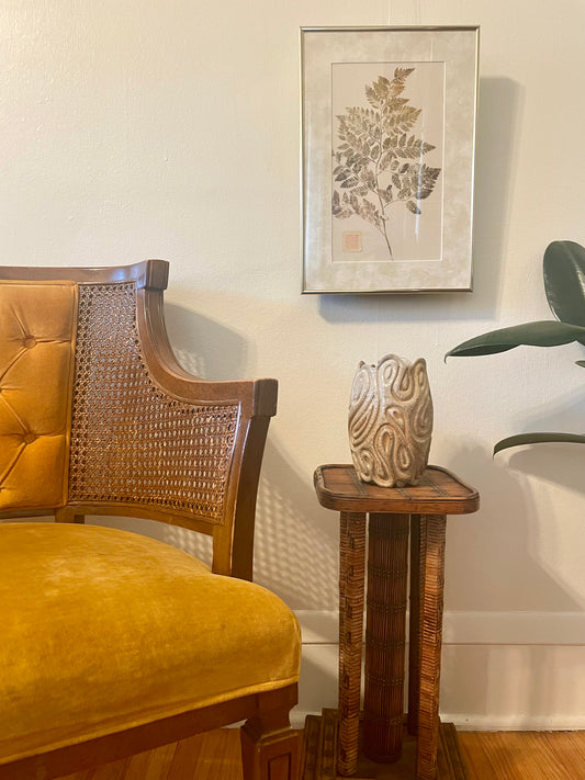 A styled interior corner featuring a mustard yellow velvet and cane armchair, a small rattan side table with a carved ceramic vase, and a framed botanical print hanging above on a cream.