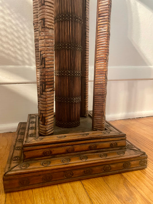 A close-up of a decorative wooden table base with rattan-wrapped legs and carved circular patterns, standing on a hardwood floor against a white wall.