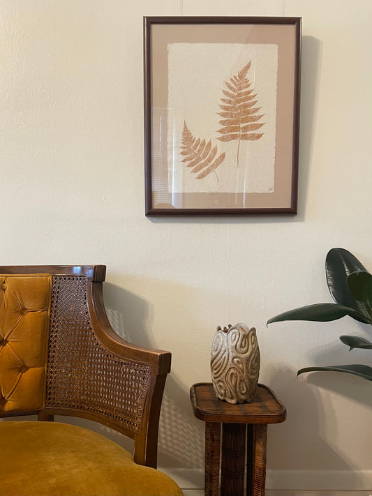 A framed botanical art print featuring two brown fern leaves on a textured beige background, styled in a warm interior setting above an armchair and a small wooden drink table with a carved ceramic vase.
