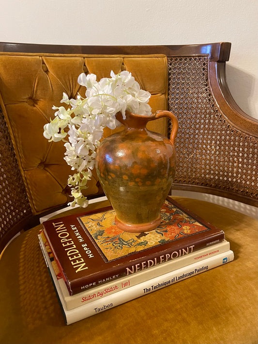 A rustic green and orange speckled ceramic vase filled with white artificial flowers, placed on a stack of three craft books on a tufted mustard-brown armchair.