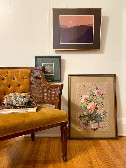 A styled interior scene showing framed wall art in a home setting. A mustard yellow tufted chair with a stack of books and a patchwork dog figurine sits on a hardwood floor, next to a framed floral painting leaning against the wall, with a framed landscape photograph depicting the silhouette of a mountain range under a pink and orange sky with a moon in the sky hanging on the wall.