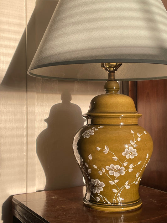 A table lamp with a tapered light blue fabric shade and a glossy olive green ceramic base decorated with white floral patterns, set on a dark wooden side table in a warm indoor setting.
