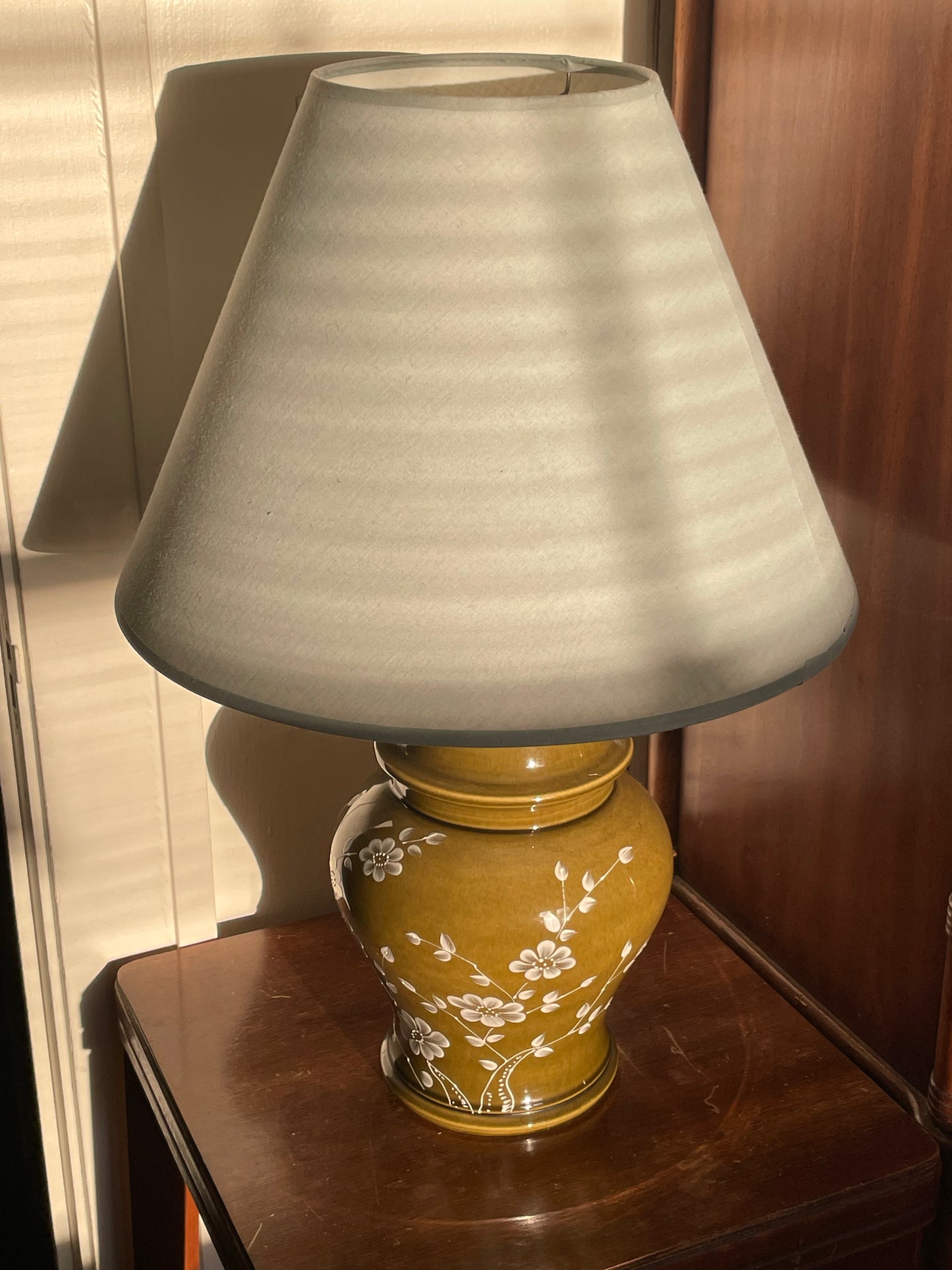 A table lamp with a tapered light blue fabric shade and a glossy olive green ceramic base decorated with white floral patterns, set on a dark wooden side table in a warm indoor setting.
