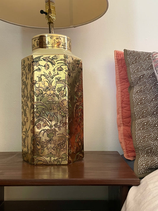 A decorative hexagonal table lamp with a gold-toned body covered in an intricate floral pattern, sitting on a dark wooden table next to a bed with patterned pillows.