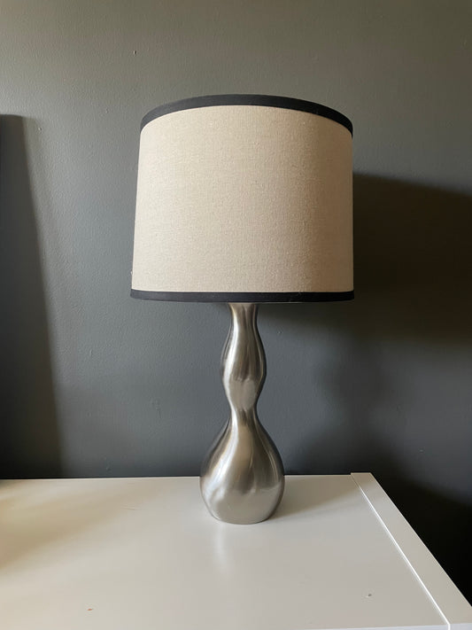 A table lamp with a textured beige fabric drum shade trimmed in black, mounted on a sculptural, hourglass-shaped metallic silver base, set on a white surface against a dark gray wall.