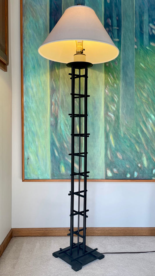 A tall black iron grid-style floor lamp with a large white conical shade, illuminated with a warm yellow glow, standing on a beige carpet in front of a large green and blue abstract painting.
