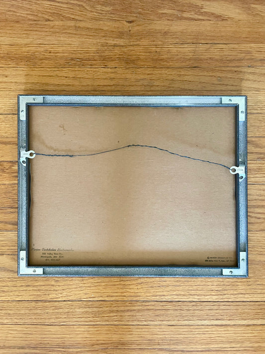 The back of a rectangular framed photo, showing a brown cardboard backing and a silver metal frame with hanging loops, placed on a wooden surface.