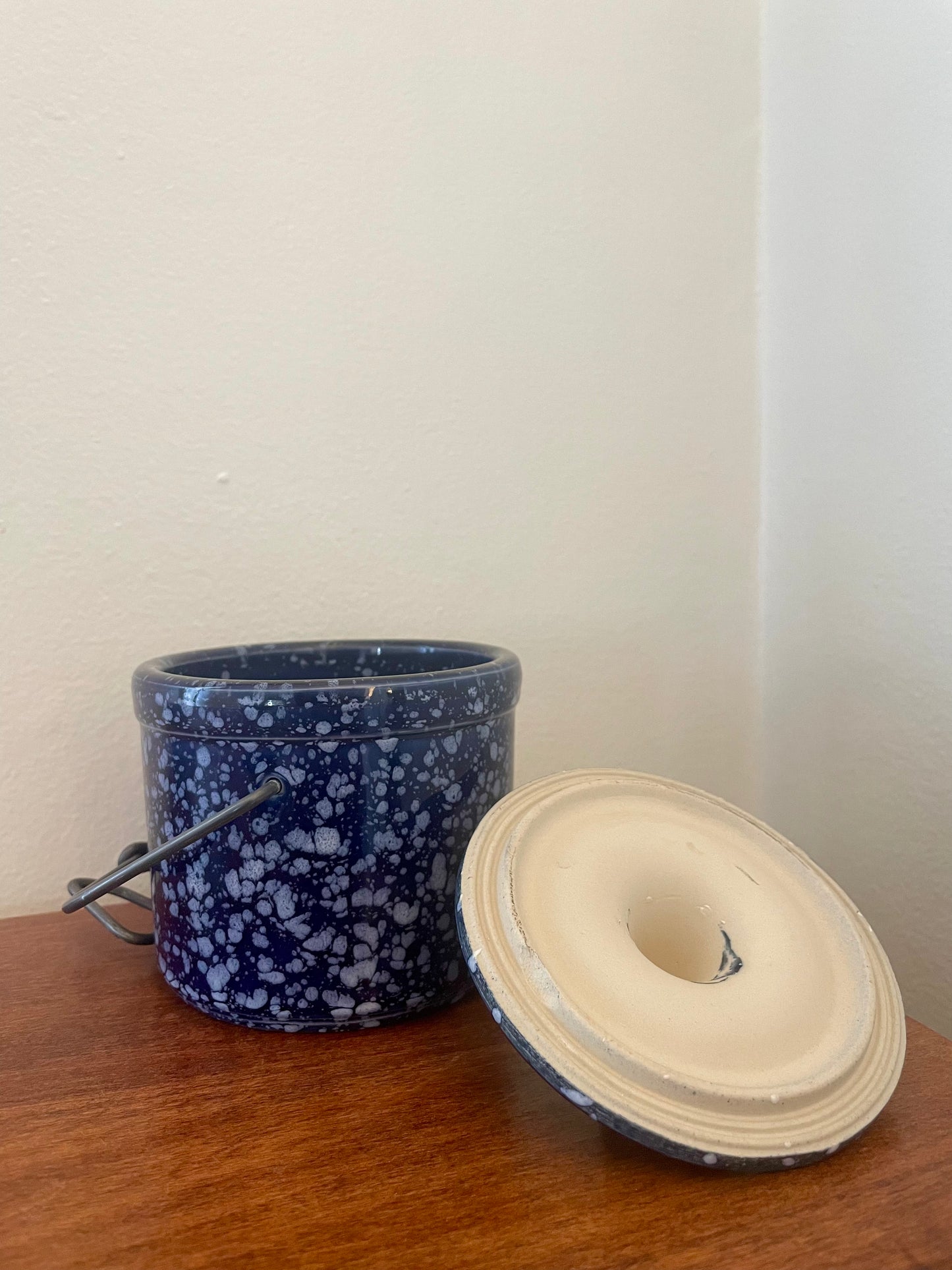 A vintage dark blue speckled stoneware canister with a metal handle, shown next to its lid sitting upside down on a wooden surface against a plain wall.