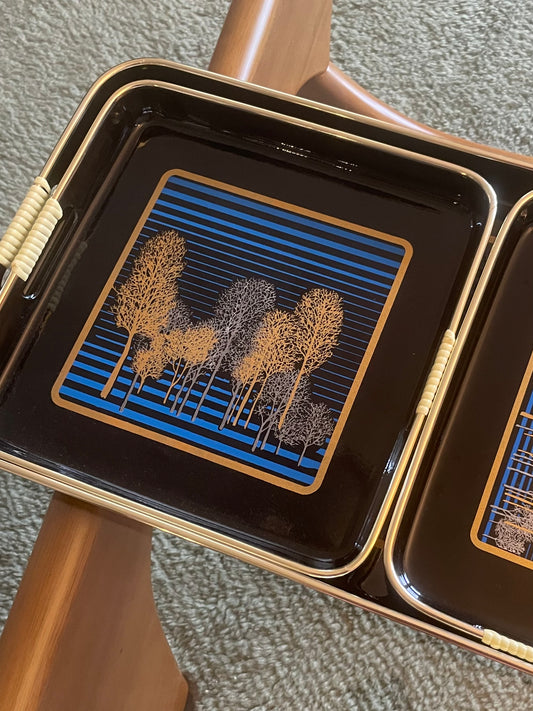 Closeup of three nested rectangular black serving trays with gold-toned edges and handles, featuring a blue striped and gold and silver tree pattern, arranged on a glass table over a textured rug.