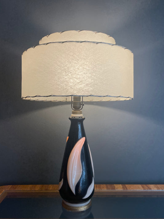 A table lamp with a textured cream-colored tiered drum shade and a glossy black painted glass base featuring a large cream leaf accent, illuminated by the bulb and from within, set against a dark gray wall.