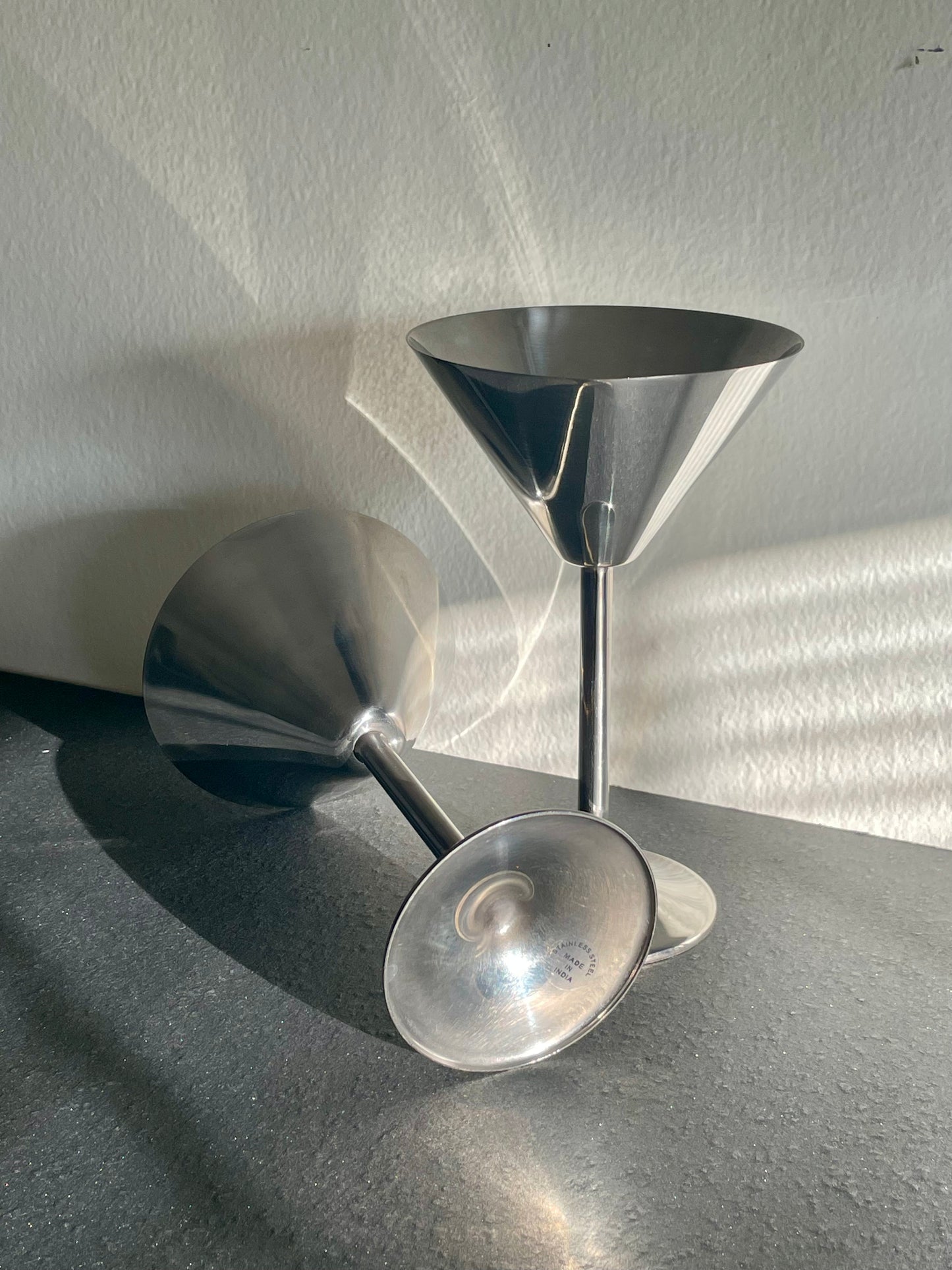 Two stainless steel martini glasses with long stems and circular conical ends, photographed on a dark tabletop against a light wall with strong sunlight and shadows.