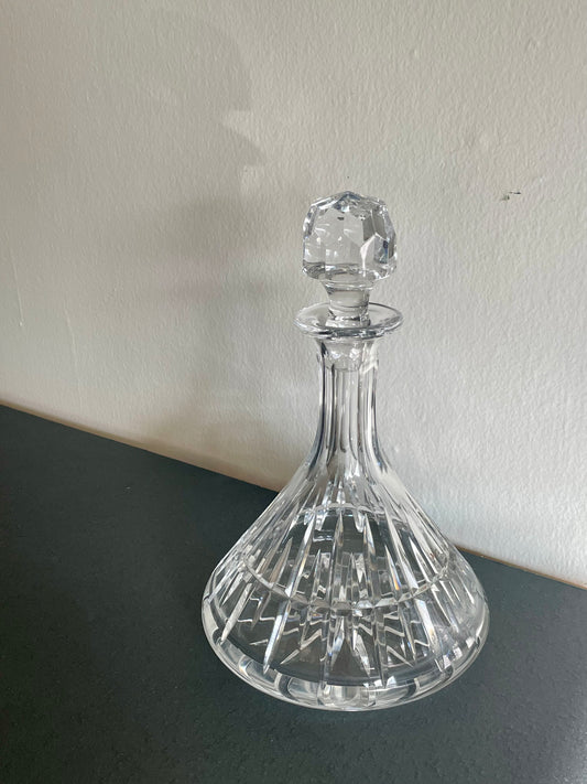 A clear cut-crystal decanter with a matching stopper, displayed on a dark surface against a light-colored wall.