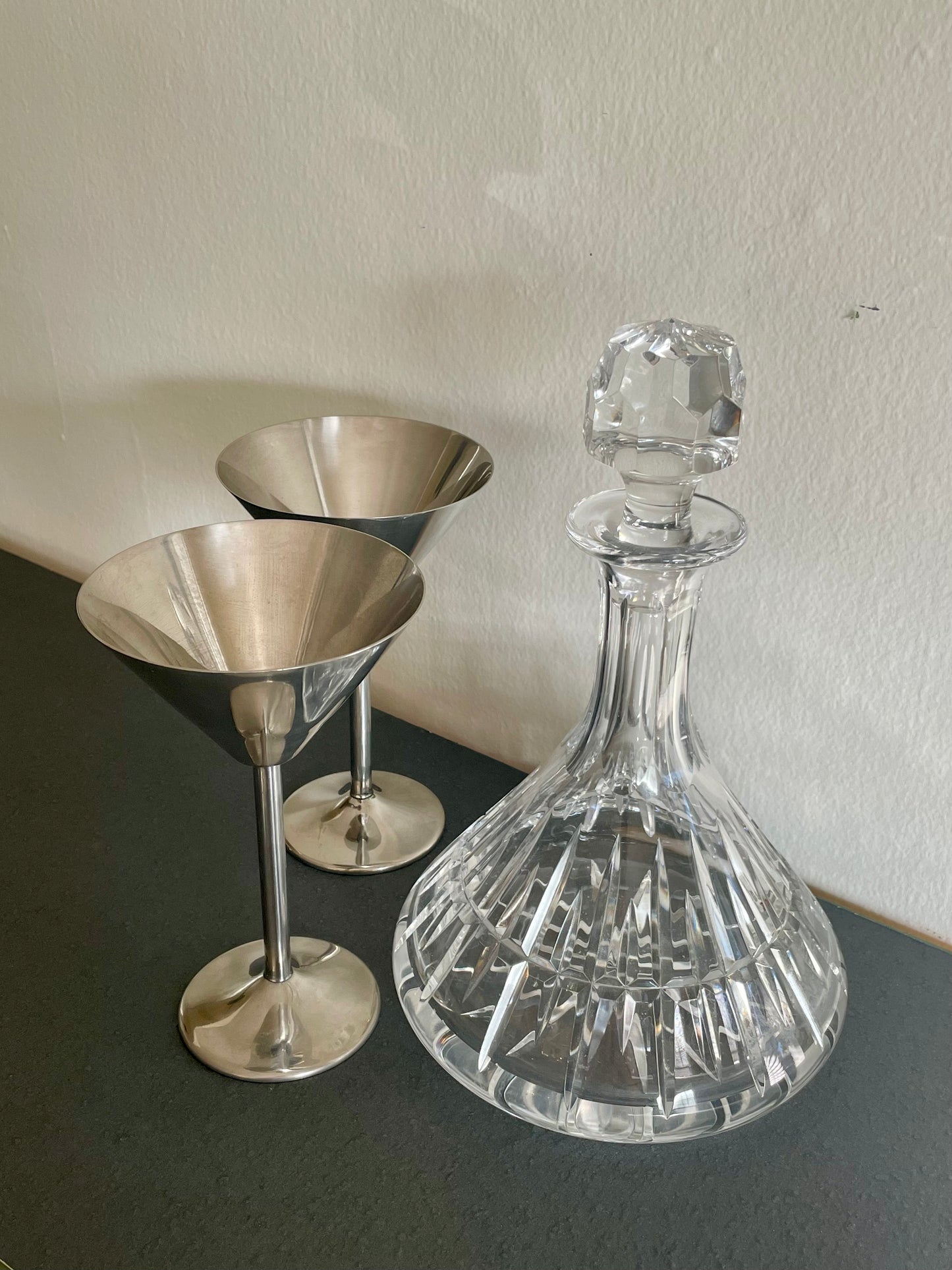 A clear cut-crystal decanter with a faceted stopper stands next to two polished silver-toned martini glasses on a dark tabletop against a light wall.