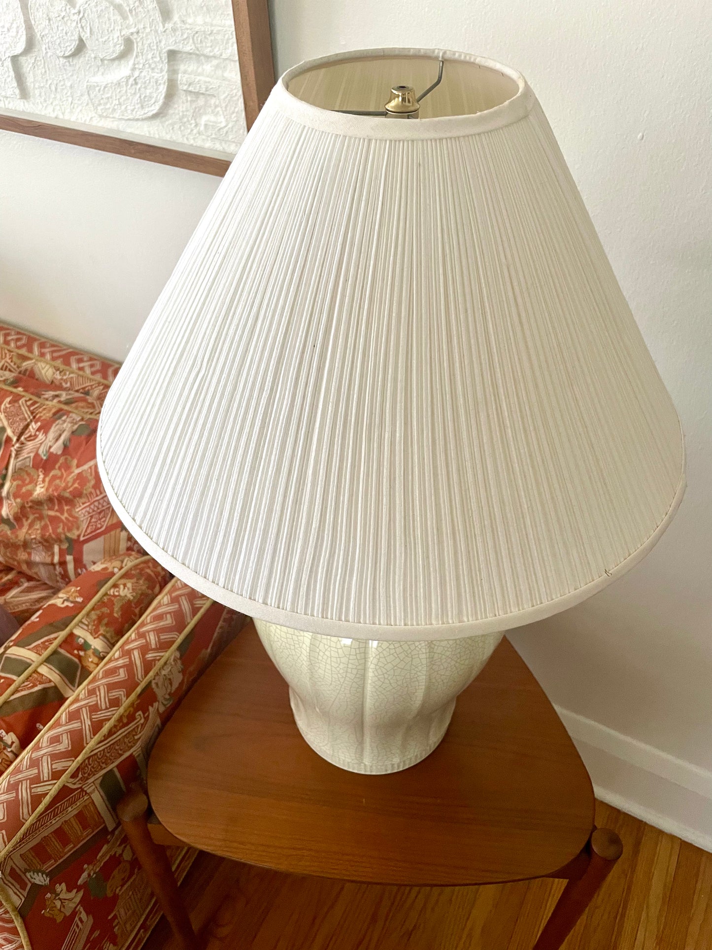 A white pleated fabric table lamp with a ribbed ceramic base, displayed on a wooden side table in a home setting next to a patterned armchair.