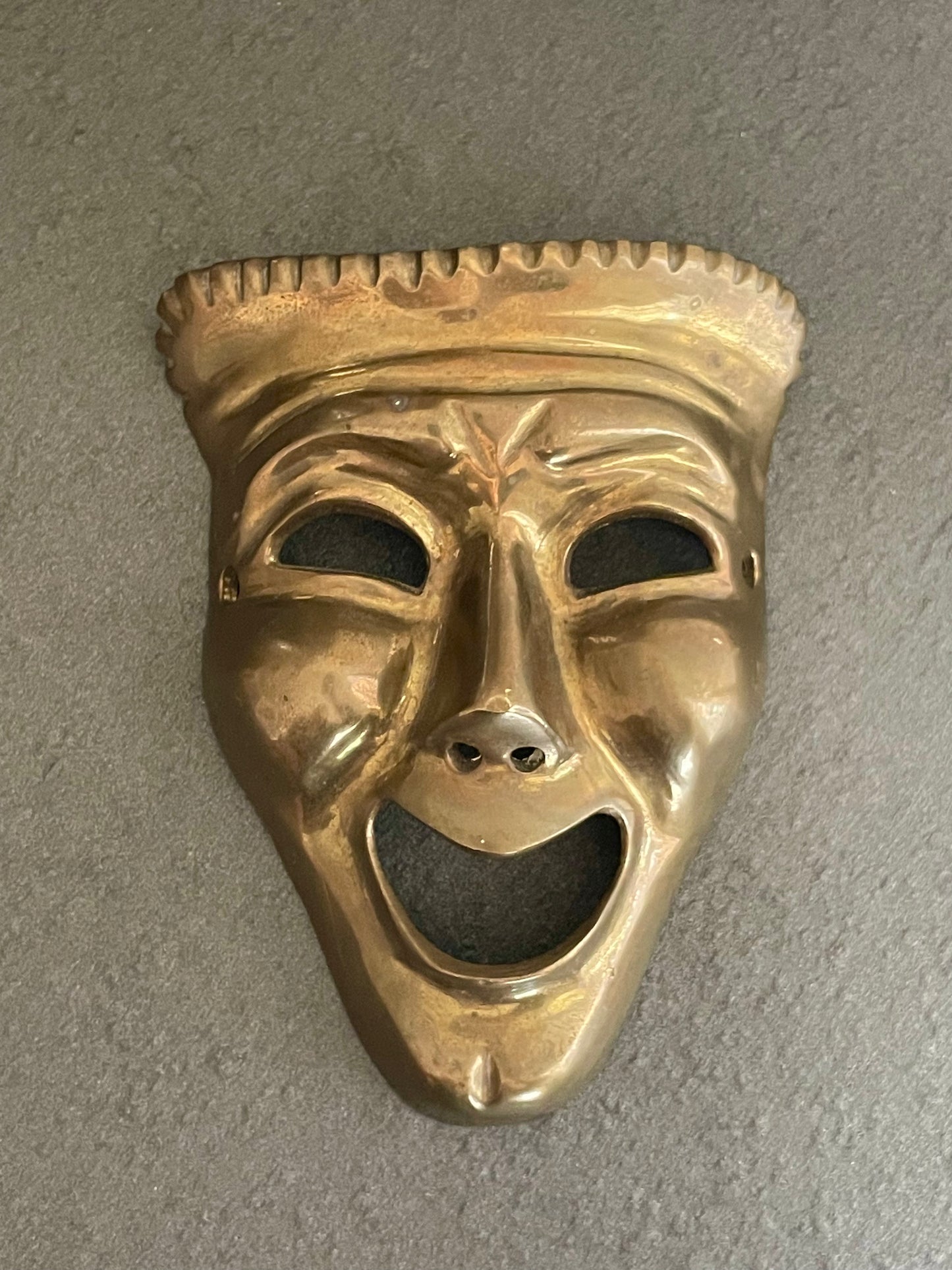A brass theatrical mask with a wide-open smiling mouth and textured top edge, photographed against a neutral gray textured background.