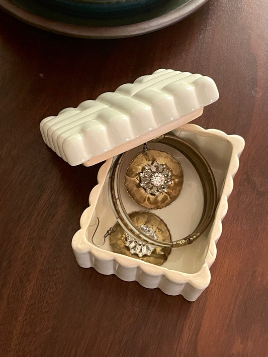 An off-white ceramic jewelry box with a scalloped edge, shown open on a dark wooden table. Inside, two gold-toned earrings with clear gemstones and a bracelet are displayed.