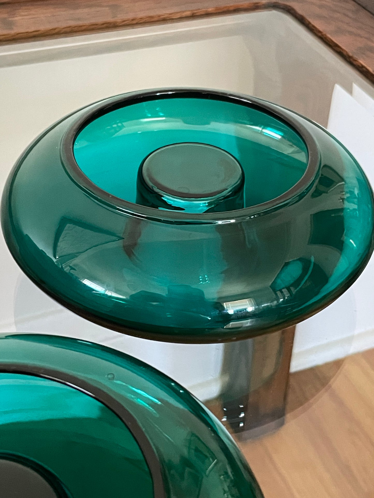 A close-up of the underside of a glossy translucent emerald green glass candleholder showing a circular opening at the base and the closed underside of the indent that would hold a candle, shown in an indoor setting with wood flooring visible.