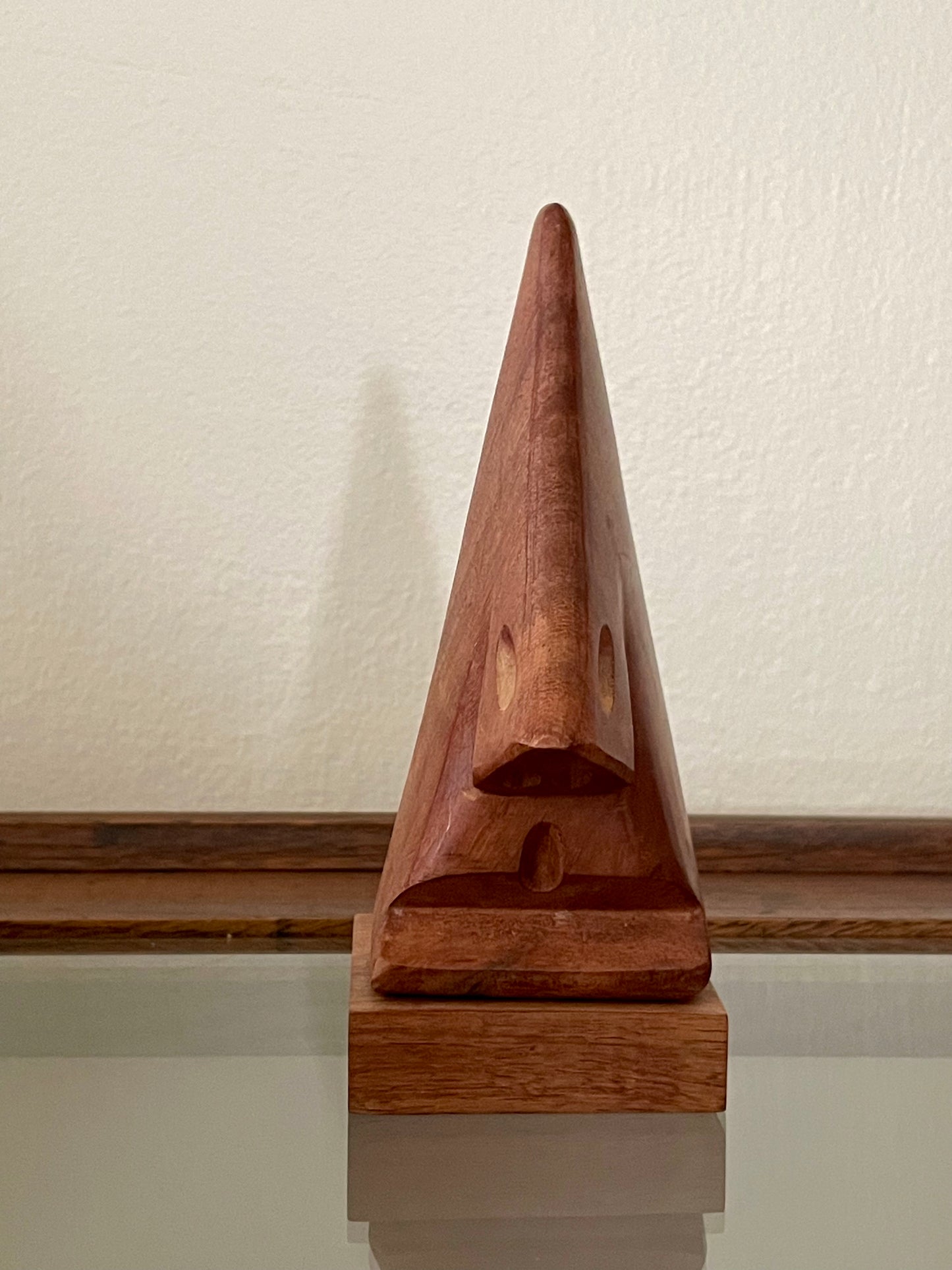 A small triangular wooden sculpture of a stylized face on a matching rectangular wooden base, photographed indoors against a light wall with a wooden ledge behind it.