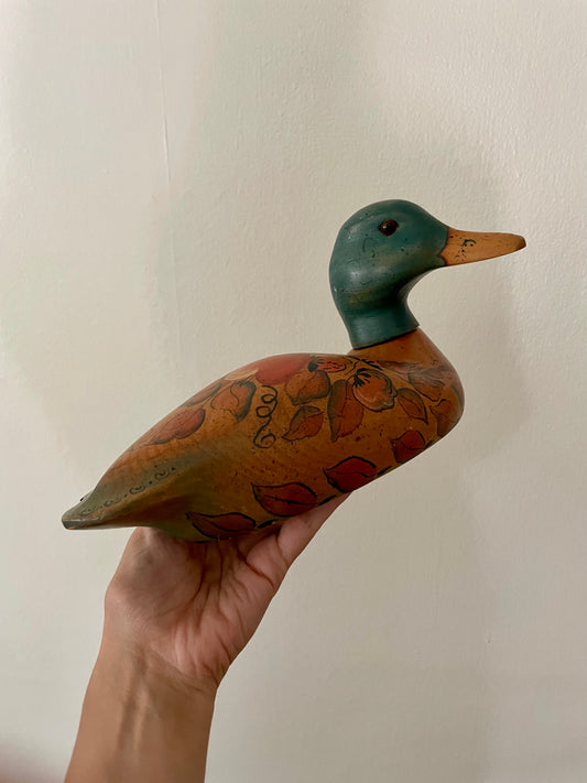 A hand holds a hand-painted wooden duck decoy against a plain off-white wall. The duck has a dark green head, a tan beak, and a brown body decorated with red and orange floral patterns and scrollwork.