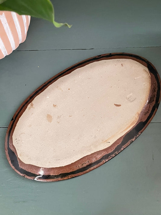 Underside of an oval ceramic serving platter with an unglazed clay center and an abstract dark brown and black glazed outer rim, shown on a painted teal wooden surface. There is a Japanese maker’s mark stamped on the bottom.