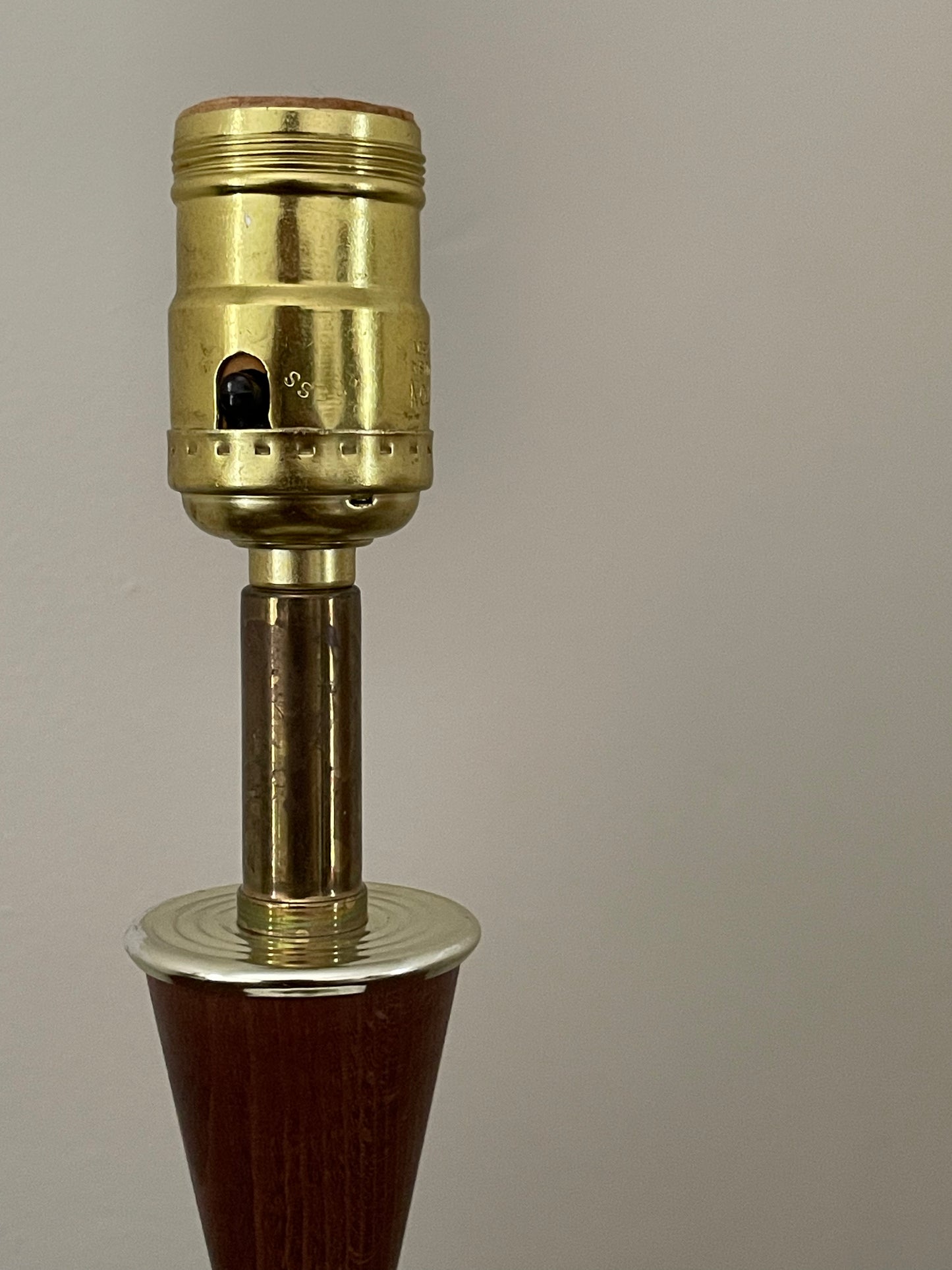 A close-up of a vintage brass lamp socket and socket cap assembly mounted on a dark brown wooden neck, set against a cream background.