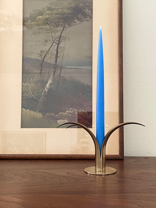 A tall, slender blue taper candle displayed in a sculptural brass holder with curved leaf-like arms, set on a wooden tabletop in front of a framed landscape painting.