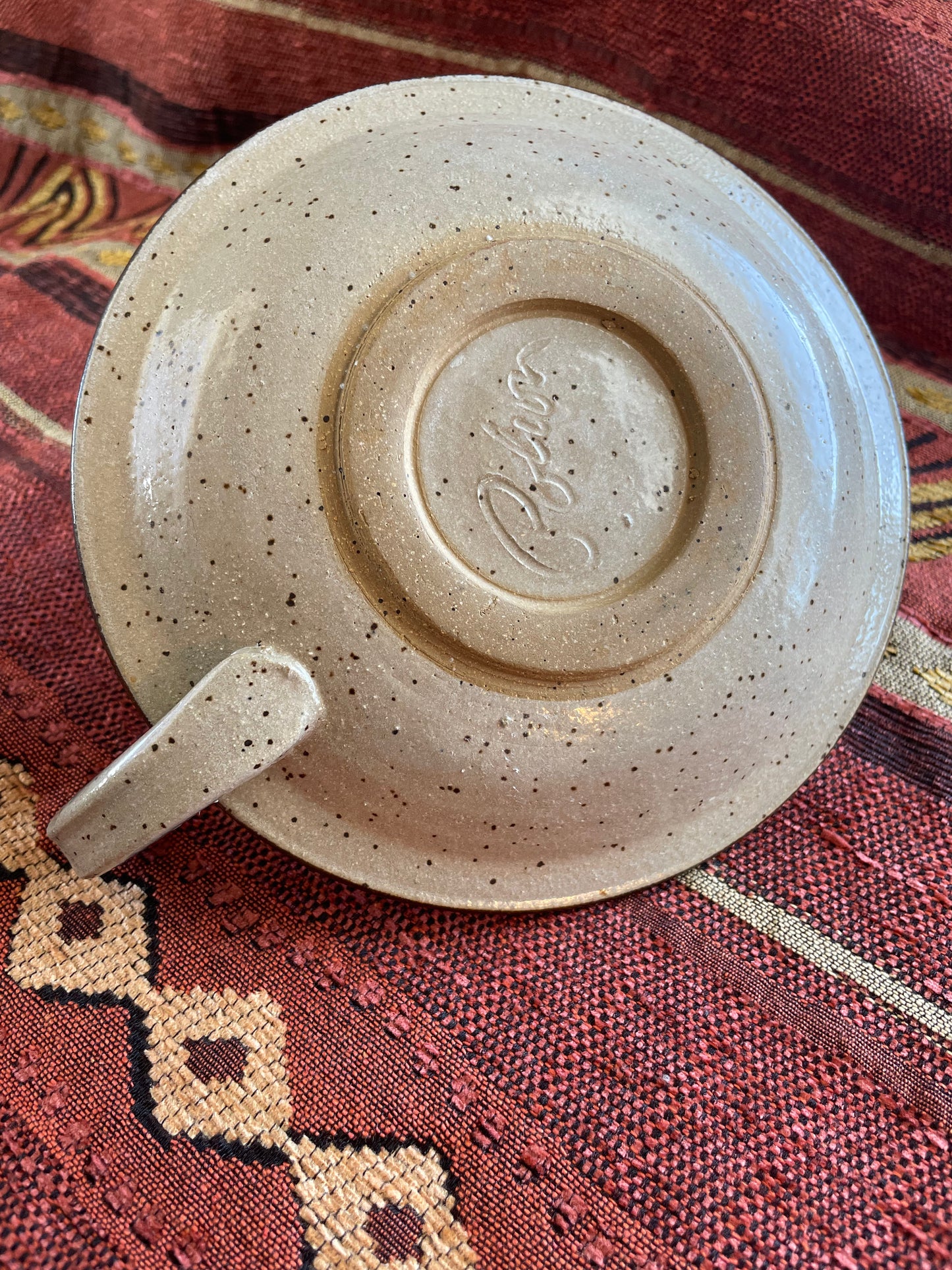 View of the underside of a circular ceramic candle holder with handle resting on a patterned fabric background.