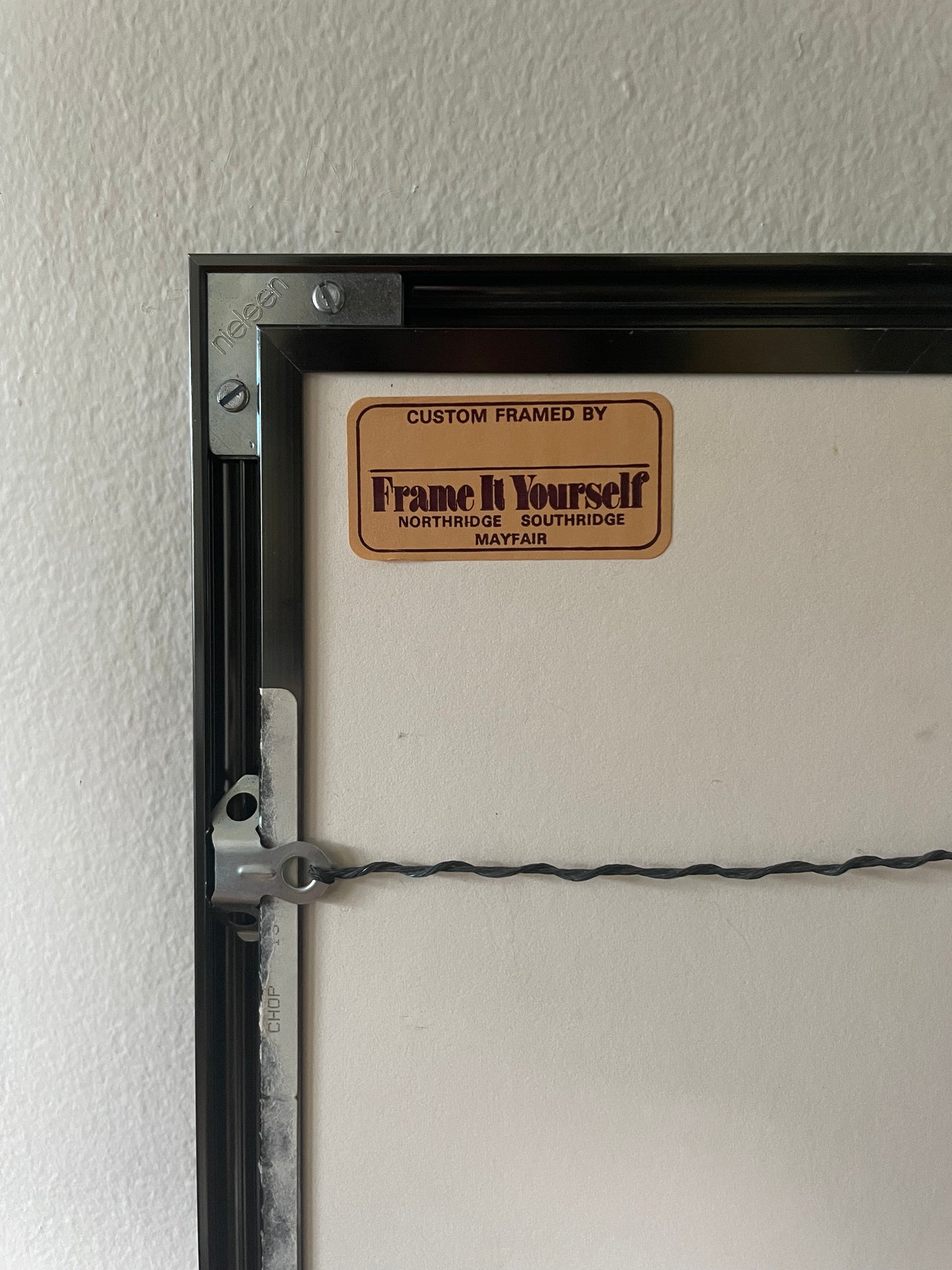 Close-up of the back side of a custom framed print, showing a black frame edge, mounting hardware, a wire support, and a tan label that reads 'Custom Framed by Frame It Yourself' on a textured off-white wall.
