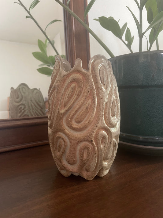 A carved ceramic sculpture in toasty beige glaze with organic, swirling patterns, displayed on a wood dresser next to a green potted plant.