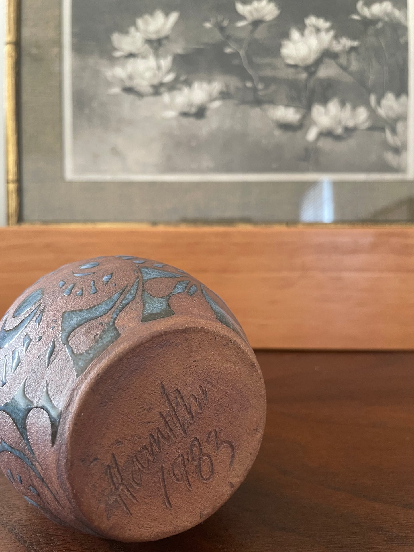 Underside of a ceramic vase with blue floral patterns and ‘Hamilton 1983' inscription resting on a wooden surface, with a framed black and white print in the background.