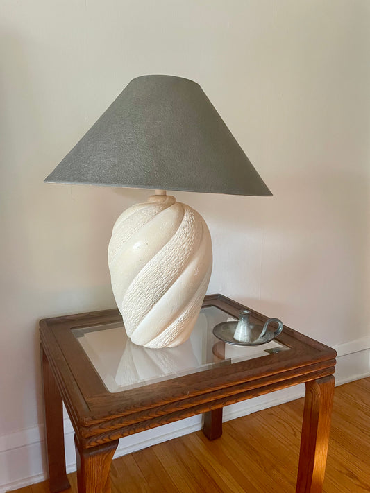 A table lamp with a textured cream-colored sculptural base and a graphite tapered shade sits on a glass-top wooden table displayed in a home setting.