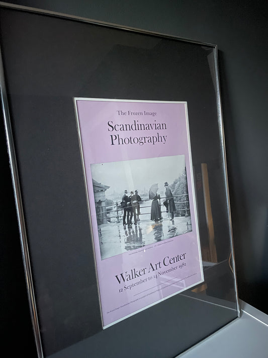 A framed lavender exhibition poster titled 'The Frozen Image: Scandinavian Photography’ for the Walker Art Center featuring a black-and-white historical photograph, shown in a silver gallery frame with grey matting.