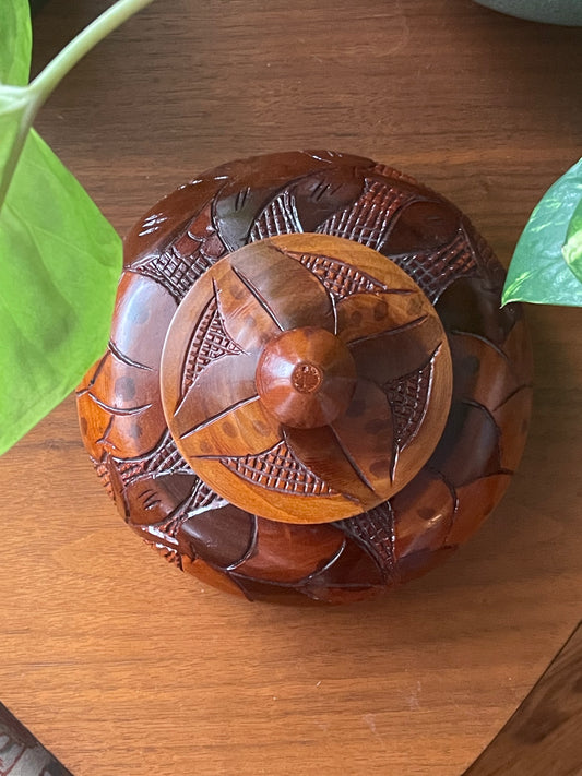 A round carved wooden box with a matching lid, featuring intricate floral and geometric engravings, placed on a wooden tabletop with green leaves visible at the edges.