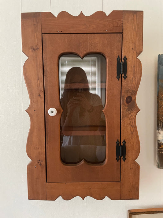 A rustic brown wooden cabinet with a scalloped door frame and a glass window door, mounted on a white wall.