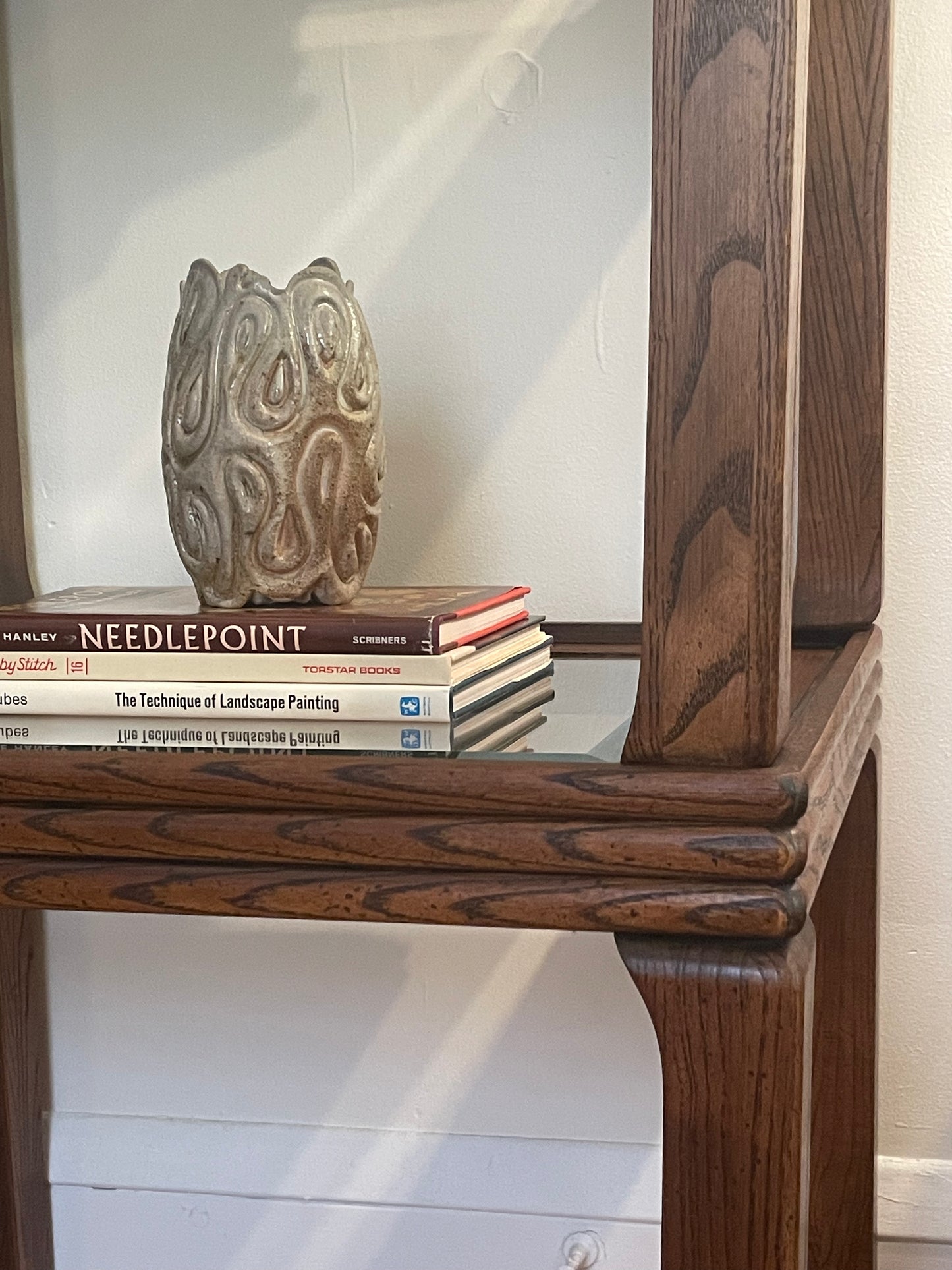 A close-up of stacked glass-top dark wood end tables, holding a decorative carved ceramic vase and a stack of books including ‘Needlepoint,’ ‘Stitch by Stitch, Vol. 16,’ and 'The Technique of Landscape Painting'.