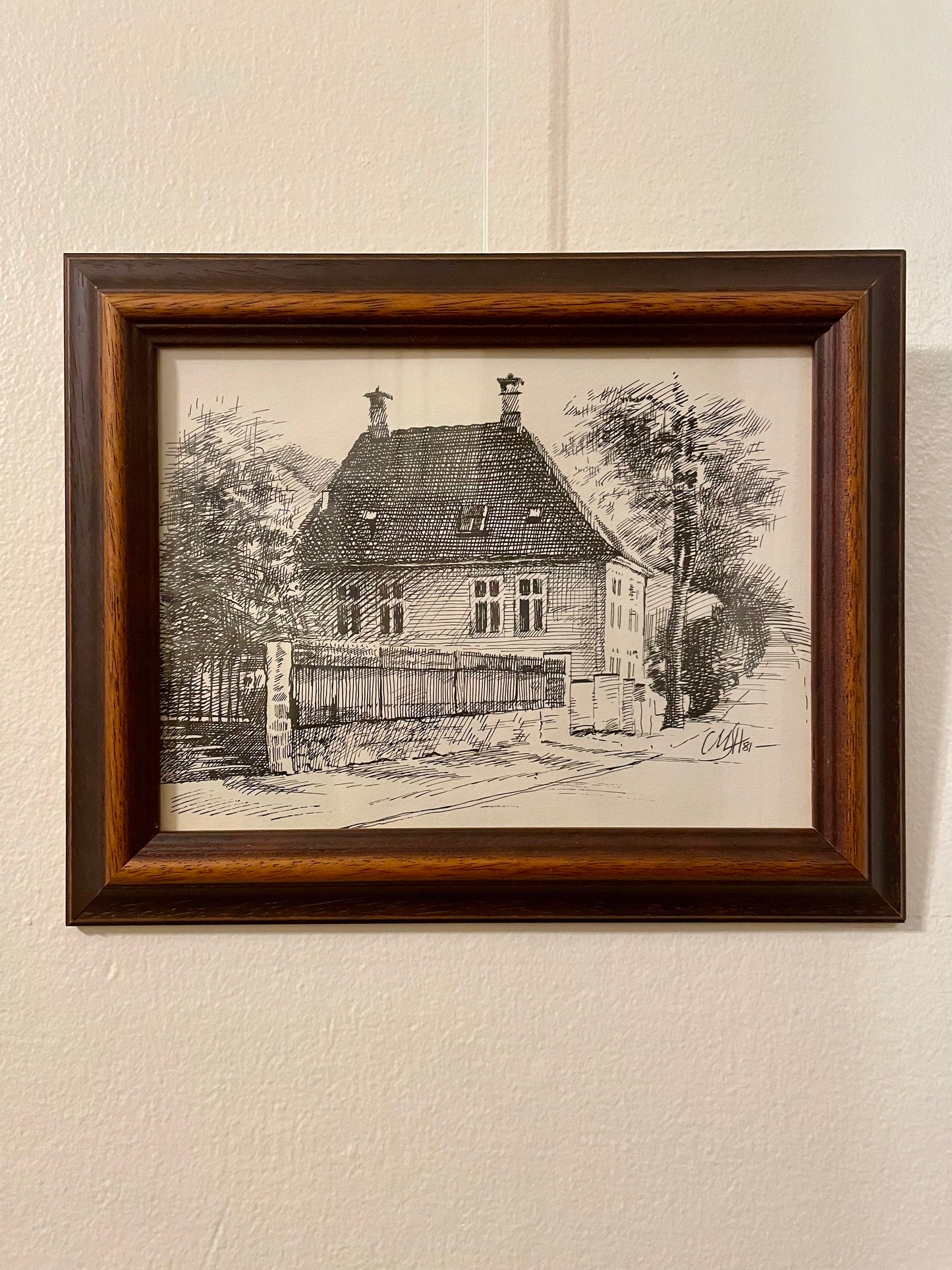 Pen and ink drawing of a house on a tree-lined street, framed in a duo-toned wood frame and mounted on a textured off-white wall.