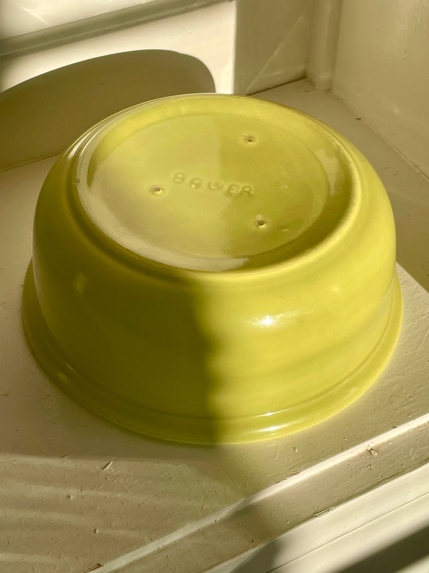 A bright yellow-green ceramic dish turned upside down on a white surface, with BAUER imprinted on the bottom. Strong sunlight casts a dark shadow across the side of the bowl.