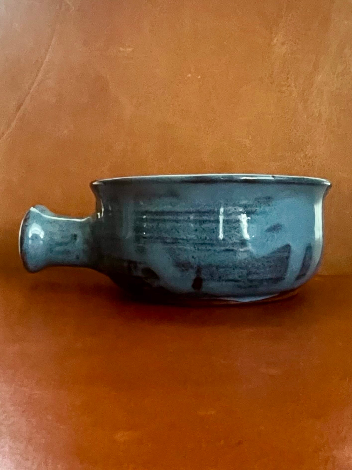 A handmade ceramic bowl with a glossy, irregular blue-gray glaze and a small side handle, set against a warm, textured brown background.