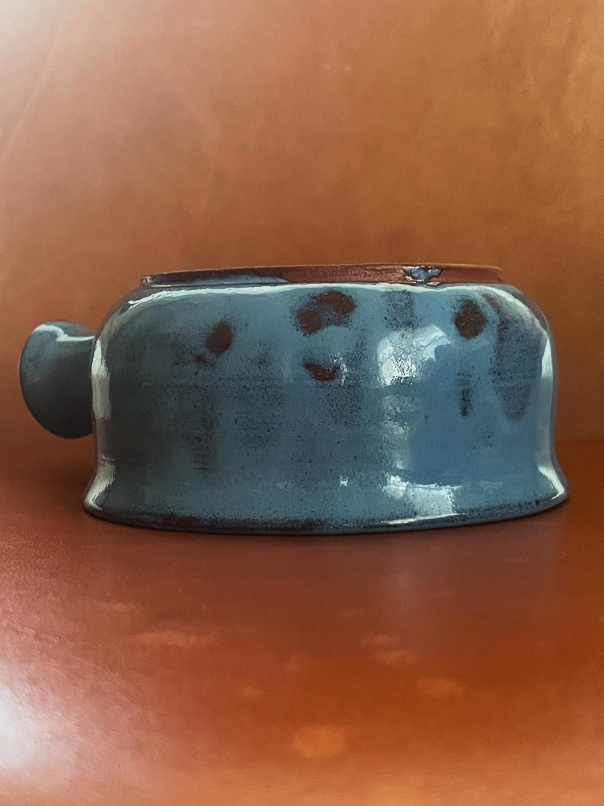 A handmade ceramic bowl with a glossy, irregular blue-gray glaze and a small side handle, sitting upside down against a warm, textured brown background.