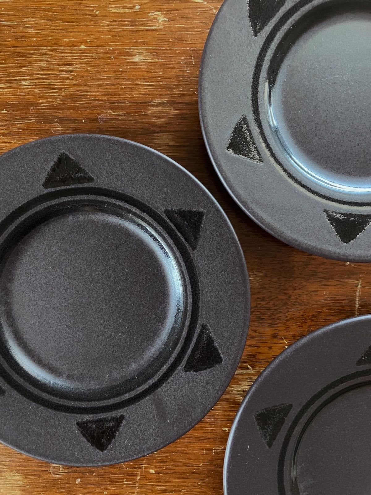 Three matte black ceramic plates with a geometric triangle pattern on the rim, arranged on a rustic wooden table.
