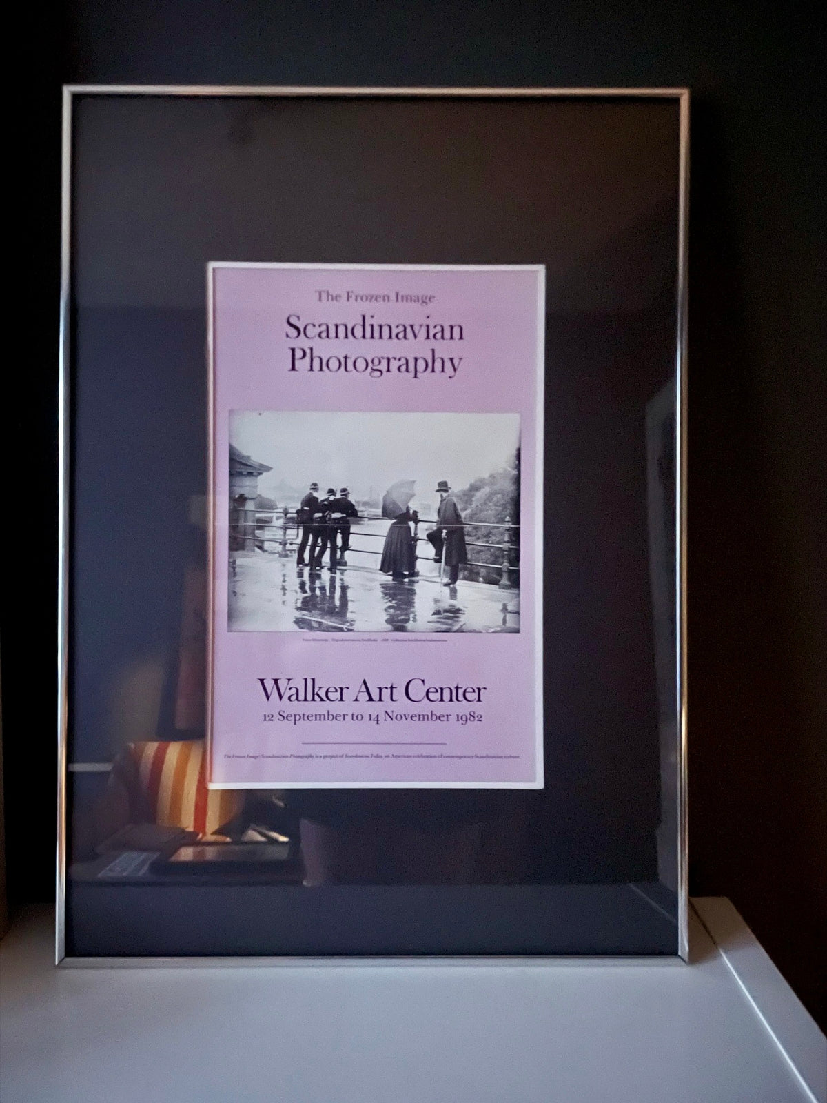 A framed 1982 exhibition poster in lavender titled 'Scandinavian Photography' from the Walker Art Center, featuring a vintage black-and-white photograph of people on a rainy street, in a silver gallery frame with graphite mat.