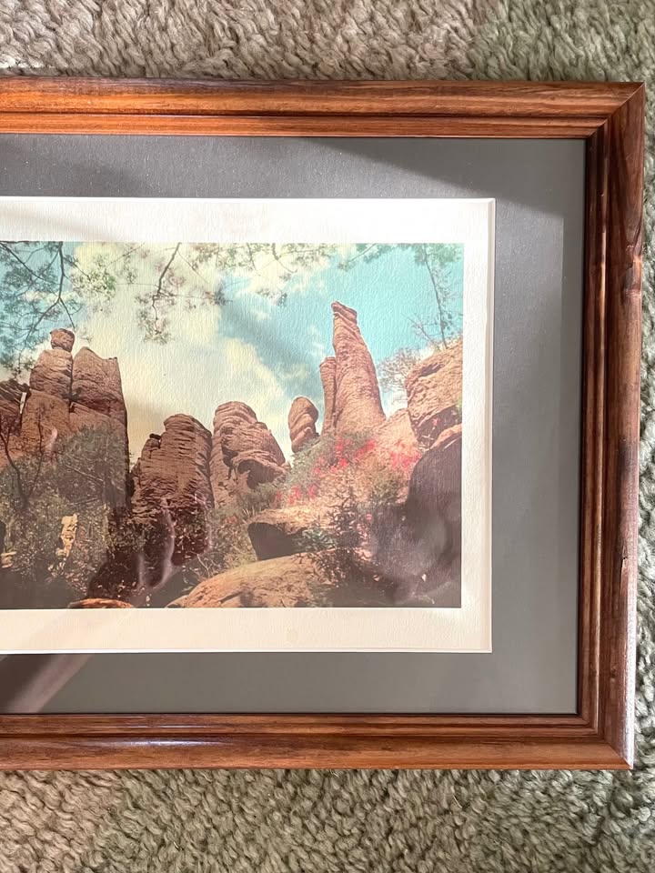 A framed art print depicting a scenic landscape with tall reddish-brown rock formations under a blue sky with a white border, matted in charcoal grey inside a brown wooden frame, resting on a textured blue-grey carpet.