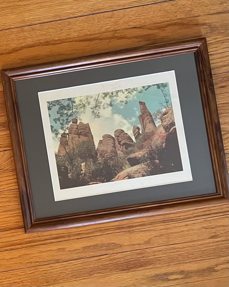 A framed art print depicting a scenic landscape with tall reddish-brown rock formations under a blue sky with a white border, matted in charcoal grey inside a brown wooden frame, resting on a wood surface.