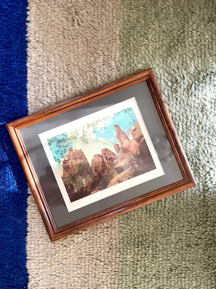 A framed art print depicting a scenic landscape with tall reddish-brown rock formations under a blue sky with a white border, matted in charcoal grey inside a brown wooden frame, resting on a textured carpet with blue, grey, and green stripes.