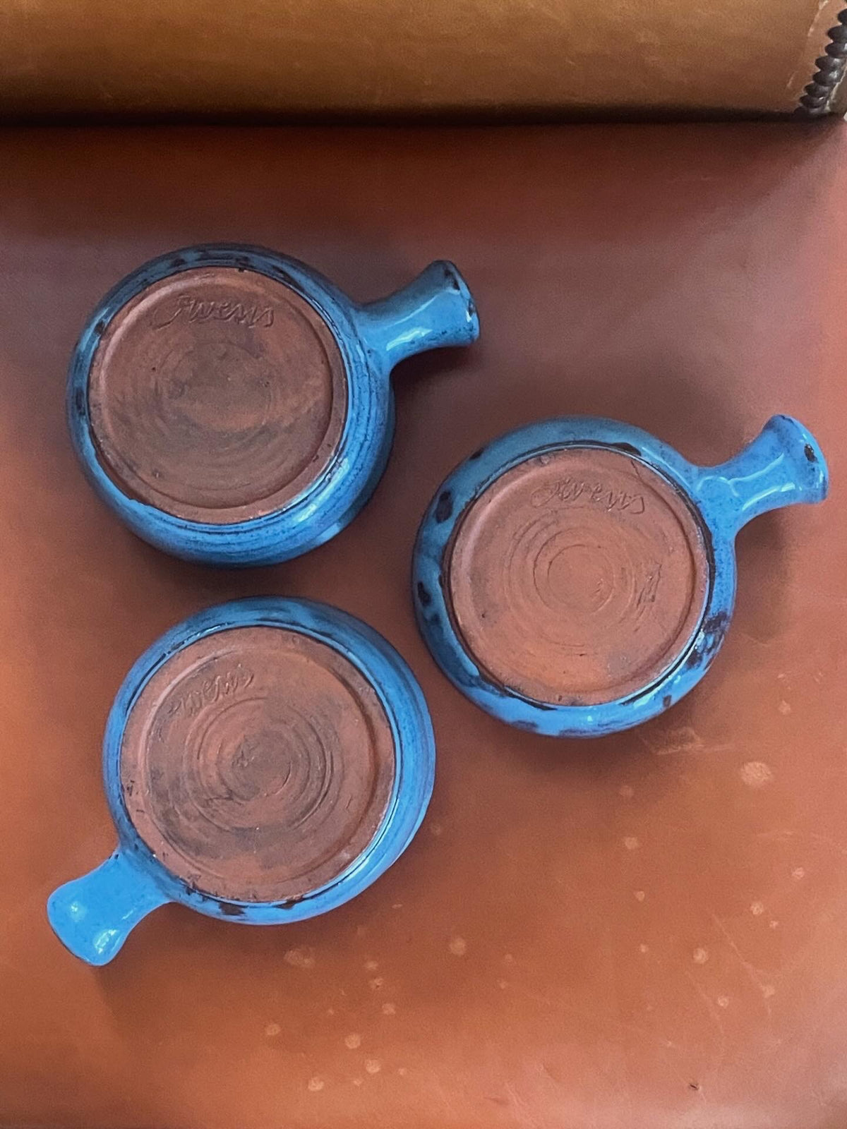 Three handmade ceramic bowls with a glossy, irregular blue-gray glaze over terracotta clay and a small side handle and signed ‘Owens', arranged upside-down on a warm brown leather surface.