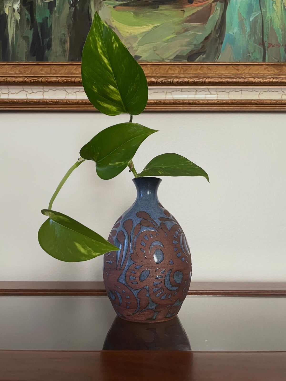 Decorative vase with a blue glazed botanical pattern over a dark unglazed clay body, with framed artwork in the background.
