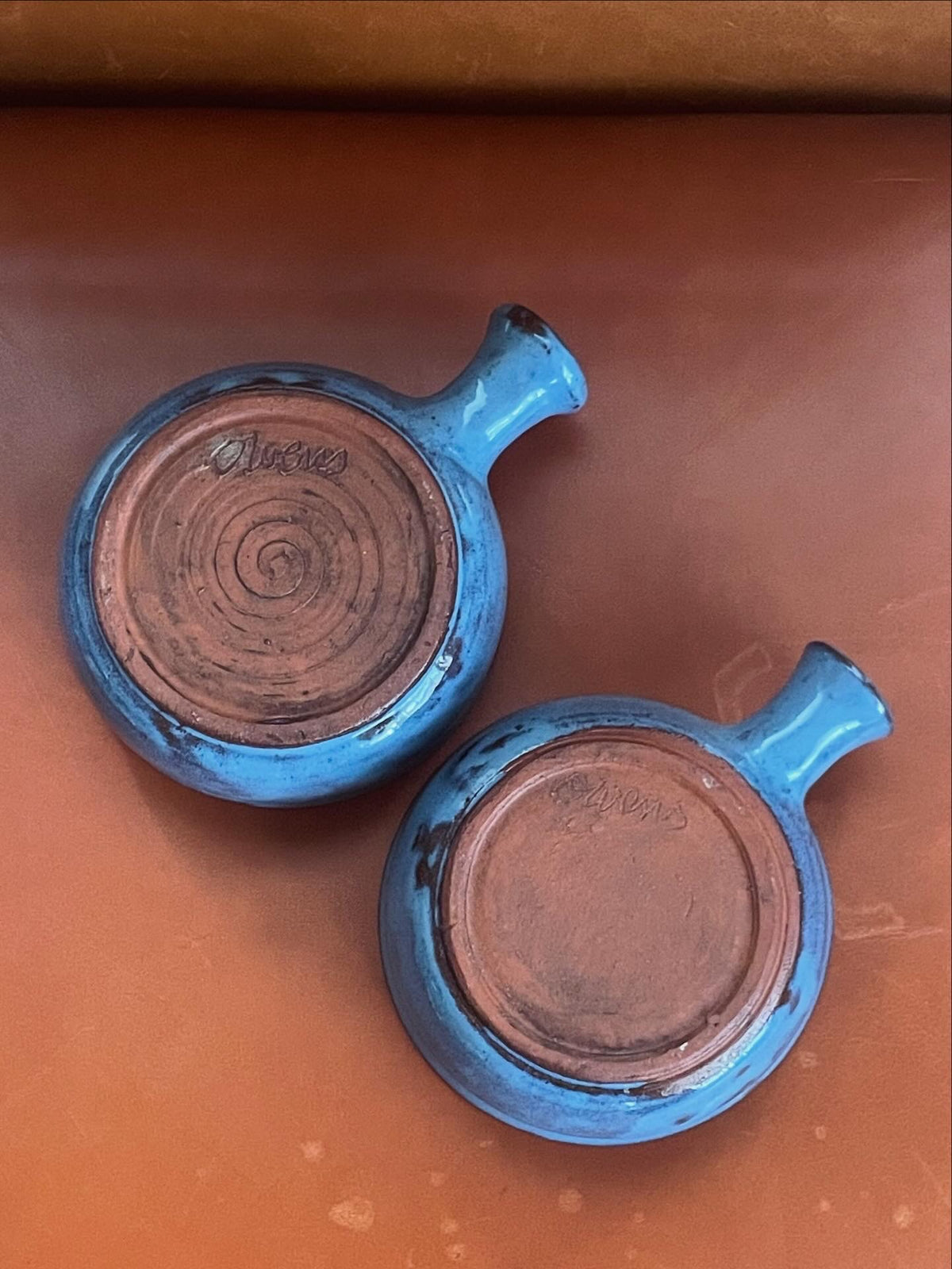 Two handmade ceramic bowls with a glossy, irregular blue-gray glaze over terracotta clay and a small side handle and signed ‘Owens', arranged upside-down on a warm brown leather surface.