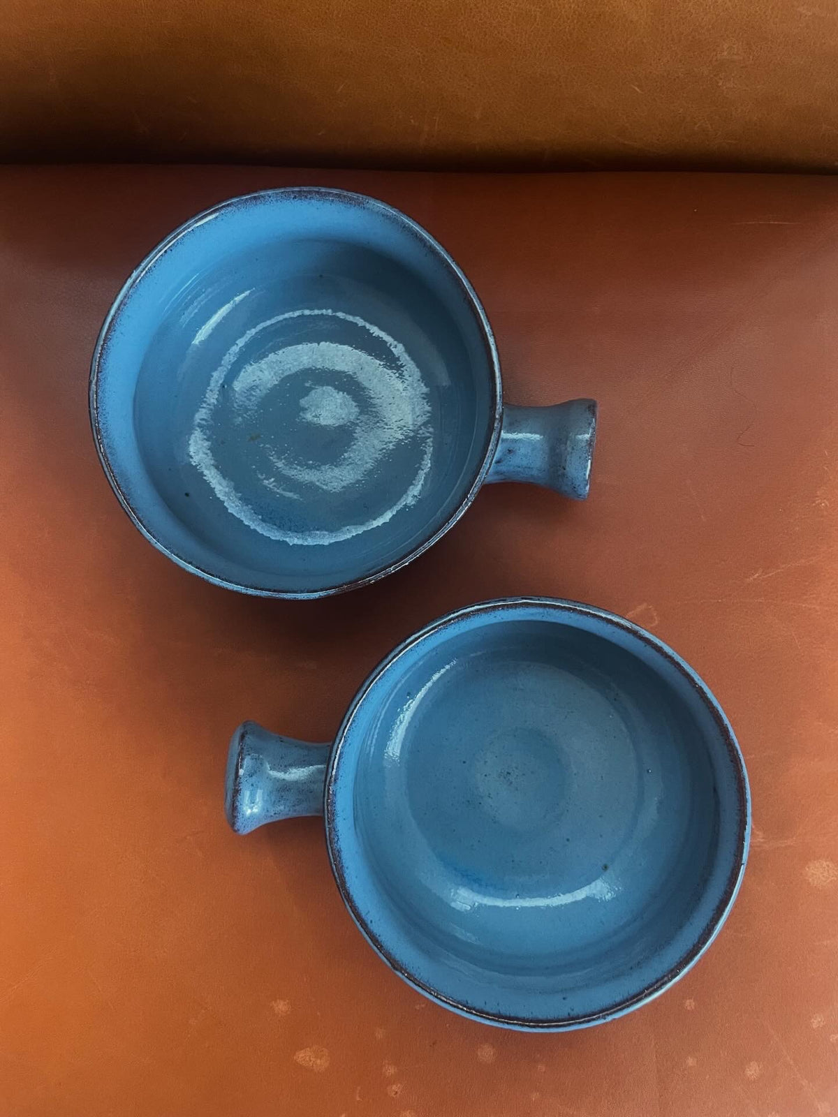 Two small blue ceramic bowls with side handles, shown from above on a brown leather surface.